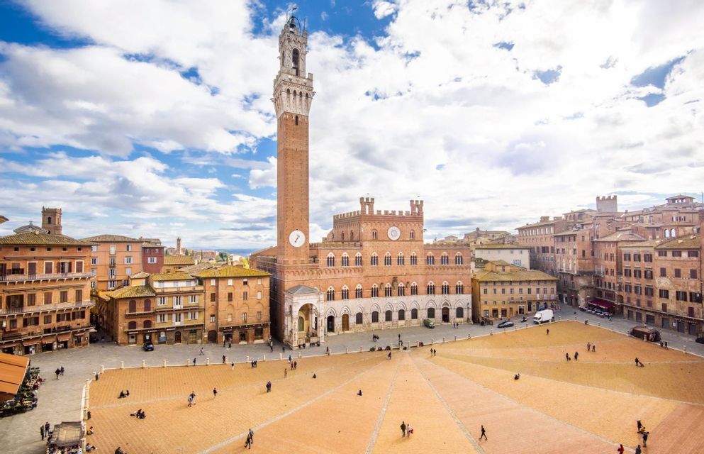 Vista elevada de una vasta y histórica plaza europea, con un alto campanario de ladrillo y personas en la plaza.