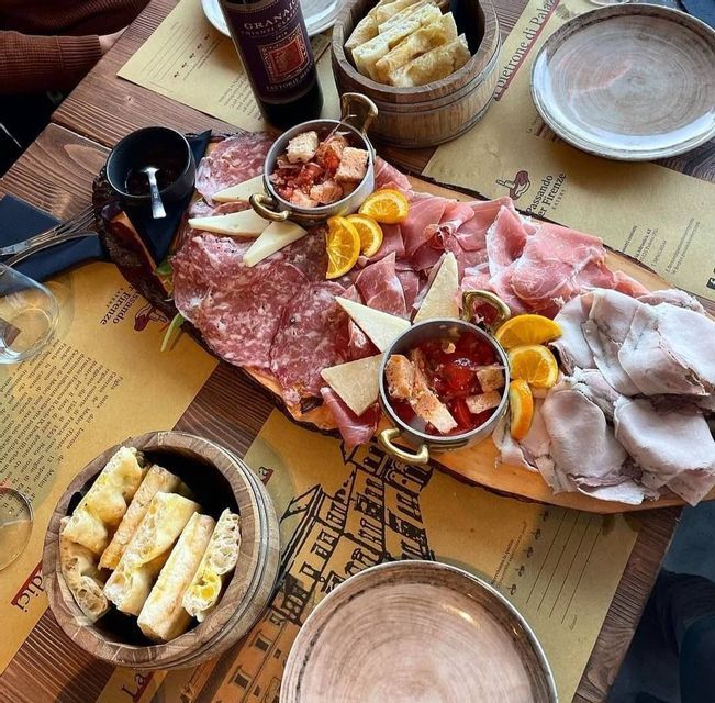 Un plato de madera con embutidos, queso, rodajas de naranja y pan servido con una botella de vino en la mesa de un restaurante.