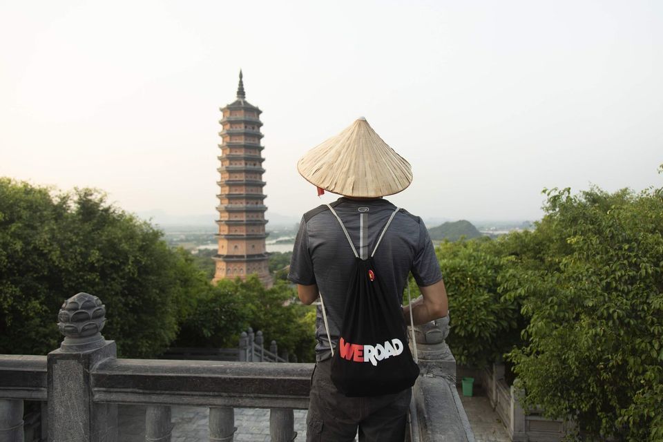 Un viaggiatore di spalle, con un cappello conico e uno zaino WeRoad, osserva una pagoda alta da un balcone di pietra.