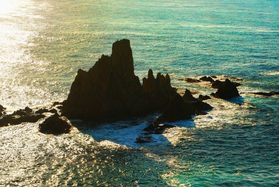 Una formación rocosa irregular en silueta se alza en el océano turquesa, mientras la luz del sol centellea sobre la superficie del agua.