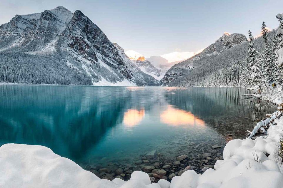Un lago calmo e limpido riflette montagne innevate e foreste di pini sotto un cielo luminoso.