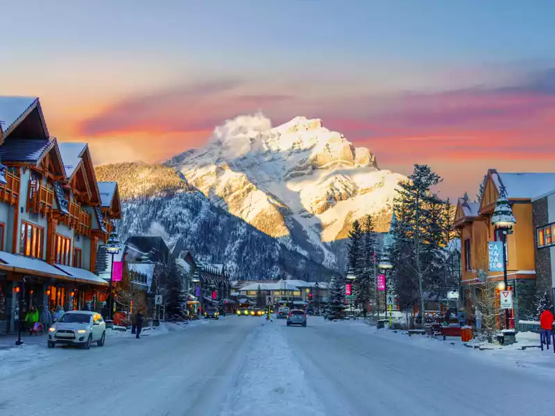 Una strada innevata in un borgo montano al tramonto, con edifici su entrambi i lati che conducono a una maestosa montagna illuminata dal sole sullo sfondo.
