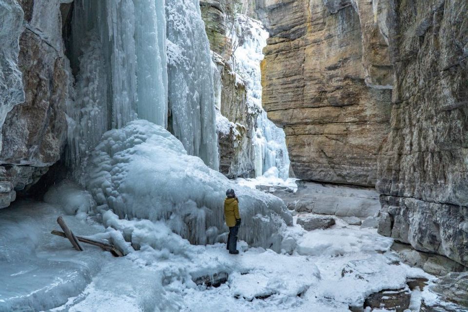 Una persona con una giacca gialla si trova in uno stretto canyon coperto di neve, guardando in alto una grande cascata ghiacciata.