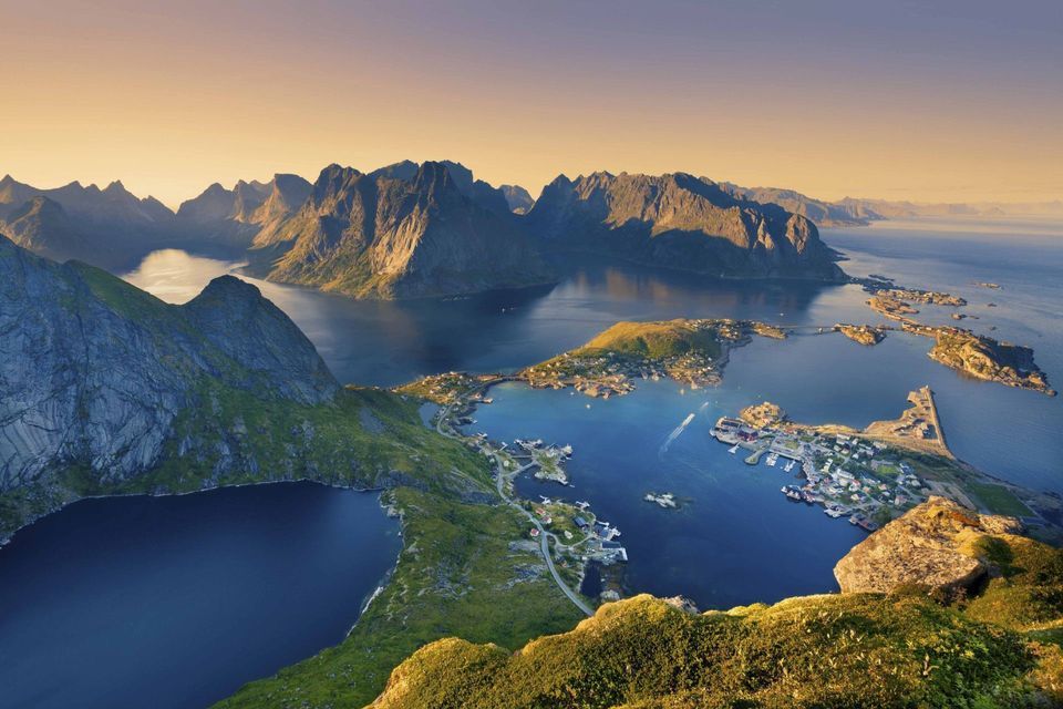 An elevated view of coastal villages on green islands surrounded by fjords and jagged mountains under a golden sky.