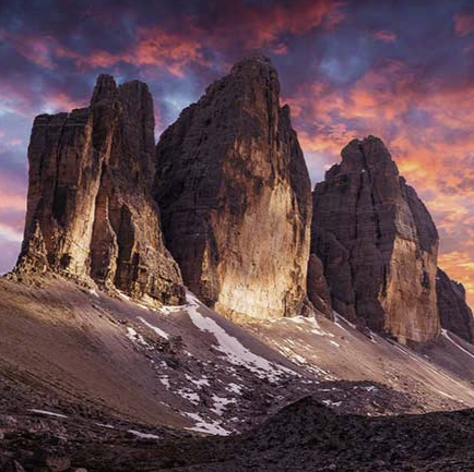 Drei zerklüftete Berggipfel mit Schneefeldern an ihren Hängen, beleuchtet von einem dramatischen und farbenprächtigen Sonnenuntergang.