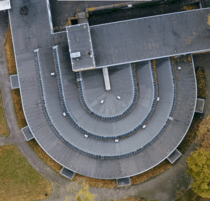 Veduta aerea dall'alto di un moderno edificio con tetto a livelli a forma di U, circondato da erba e foglie autunnali.