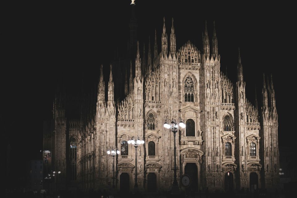 The intricate facade of a large Gothic cathedral with many spires, illuminated by lights against a dark night sky.
