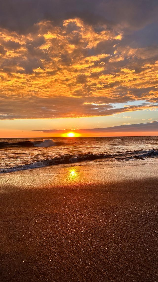 Le soleil se couche sur l'océan, projetant un reflet doré sur l'eau et la plage de sable humide sous un ciel nuageux.