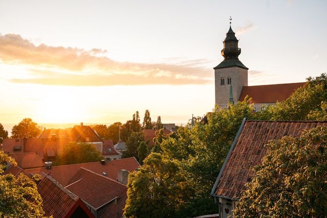 Ein steinerner Kirchturm erhebt sich während eines goldenen Sonnenuntergangs über die roten Ziegeldächer und grünen Bäume einer Stadt.