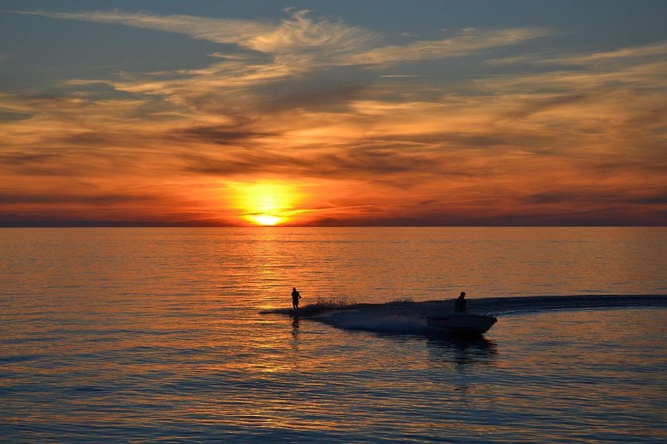 Eine Person beim Wasserski auf dem Meer, von einem Motorboot gezogen, vor einem leuchtend orangefarbenen Sonnenuntergang.