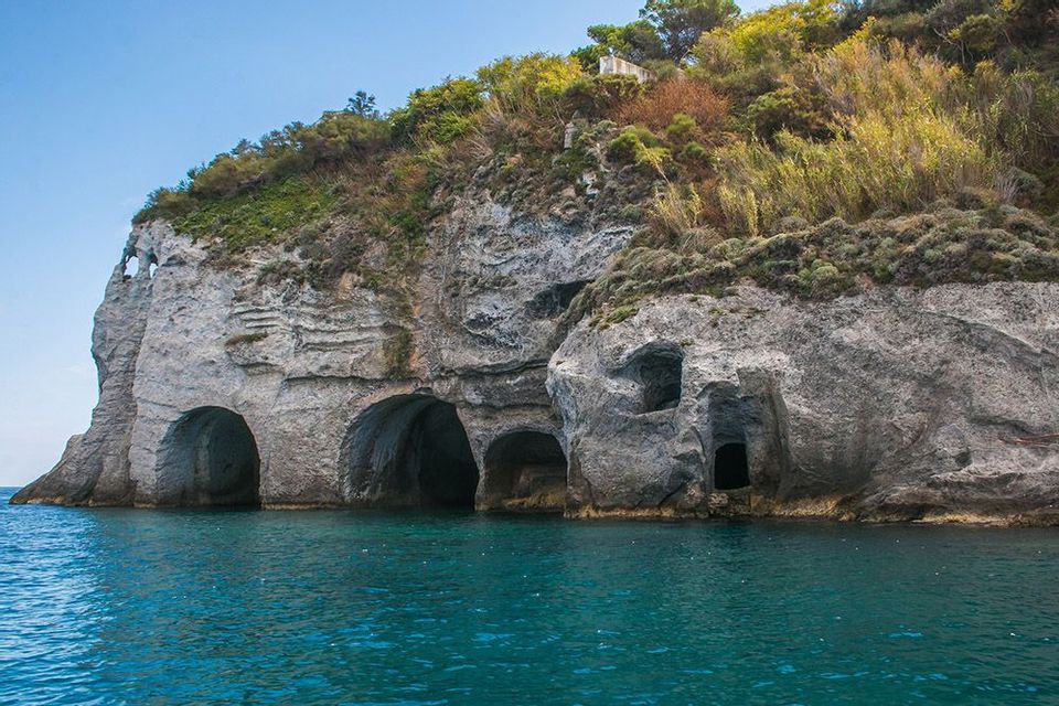 Una scogliera rocciosa con diverse grotte marine alla base, ricoperta di vegetazione verde e gialla, che emerge da acque turchesi.