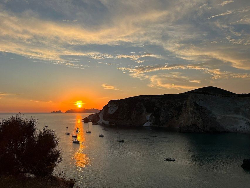 Il sole tramonta su un mare calmo, illuminando una baia dove diverse barche a vela sono ancorate vicino a scogliere rocciose e scure.