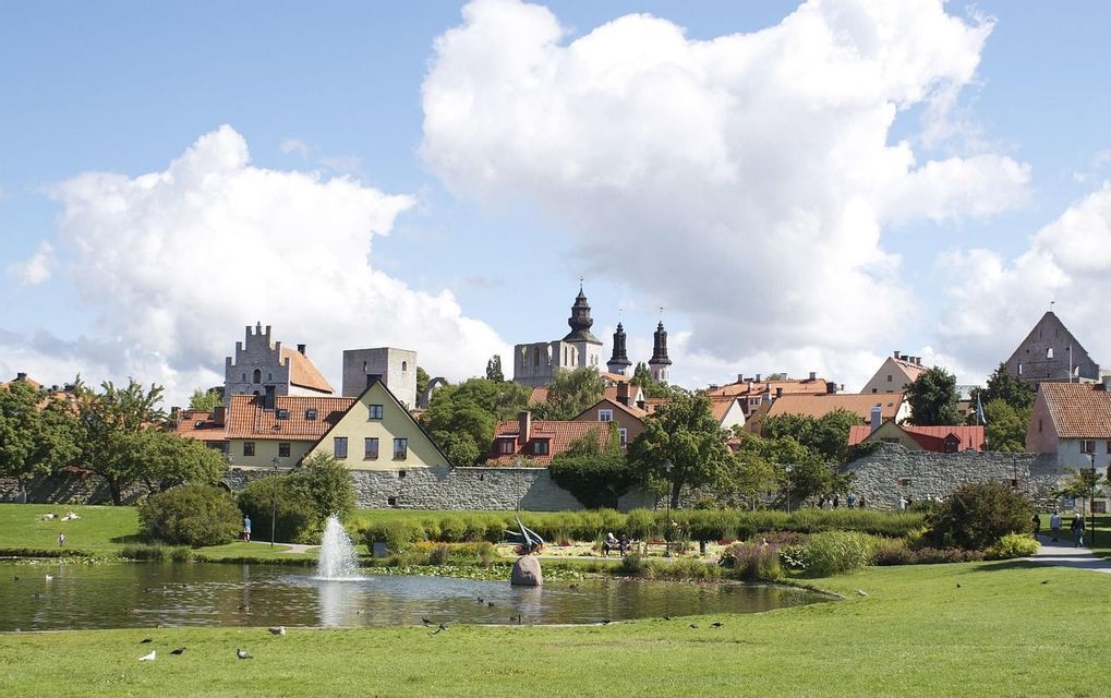 Eine historische Stadt mit Steintürmen und rot gedeckten Häusern, gesehen von einem grünen Park mit Springbrunnen in einem Teich.