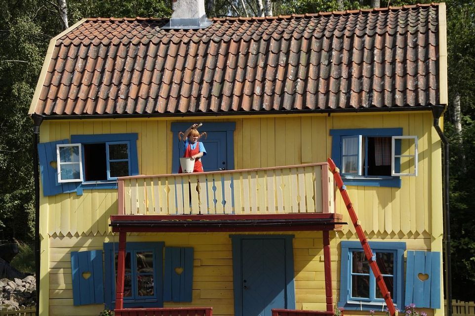 Eine Schaufensterpuppe von Pippi Langstrumpf steht auf dem Balkon eines gelben Hauses und hält einen weißen Eimer und einen Pinsel.