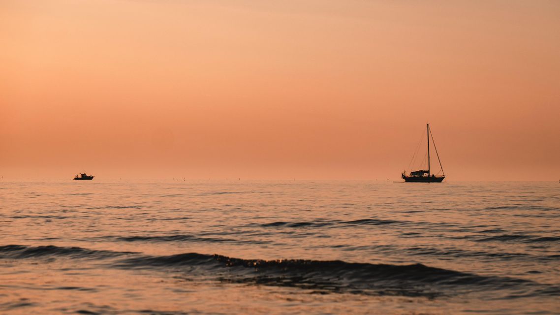 Una barca a vela in silhouette e un'altra imbarcazione galleggiano sul mare calmo sotto un cielo arancione e velato al tramonto.