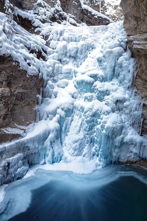 Una grande cascata ghiacciata e innevata scende da un canyon roccioso in una pozza d'acqua scura e gelida sottostante.