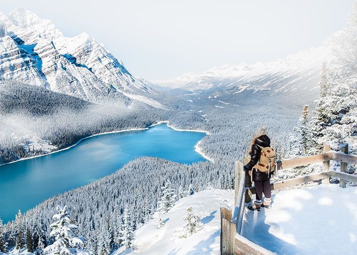 Un escursionista con uno zaino si trova su un punto panoramico innevato che domina una valle con un lago blu, foreste innevate e montagne.