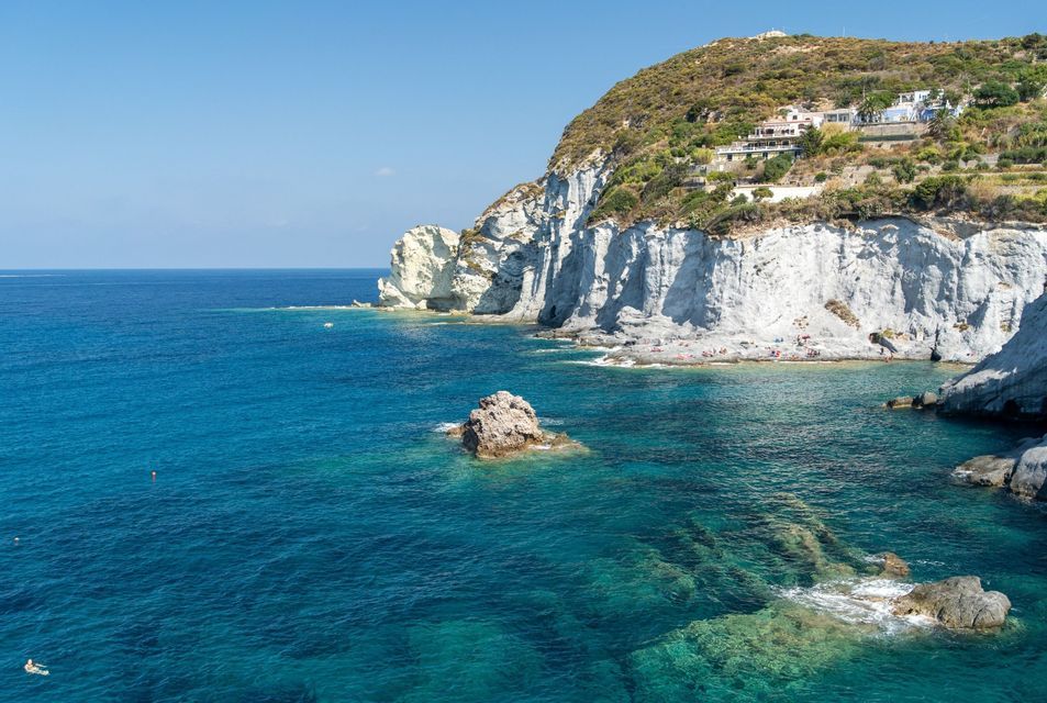 Vista dall'alto di una cala assolata con scogliere bianche e una collina verde, dove acque turchesi limpide lambiscono una piccola spiaggia con persone.