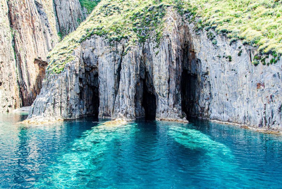 Diverse grotte marine scure alla base di una scogliera rocciosa con la cima erbosa, che si affacciano su acque limpide e turchesi.