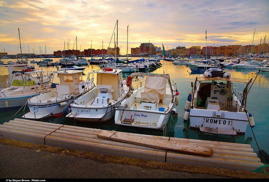 Molte barche sono ormeggiate in un porto tranquillo al tramonto, con edifici colorati sul lungomare sullo sfondo.