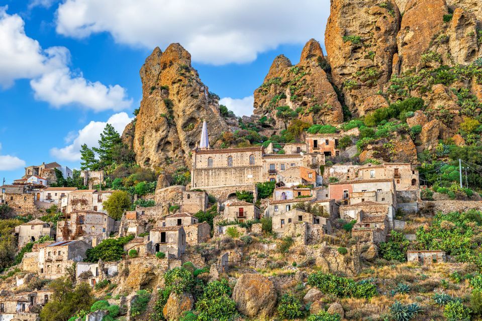 Un borgo storico con case in pietra e una chiesa incastonata alla base di formazioni rocciose frastagliate sotto un cielo parzialmente nuvoloso.