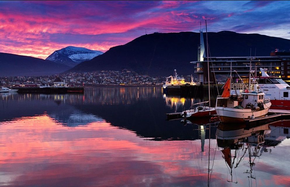 Un porto con barche ormeggiate riflette un cielo vibrante rosa e viola al tramonto, con una città di montagna illuminata sullo sfondo.