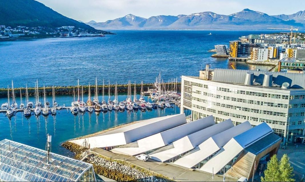 Una vista aérea de un puerto lleno de veleros, flanqueado por edificios modernos y una ciudad con montañas nevadas al fondo.