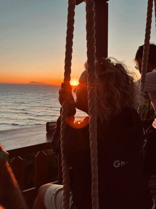 A person on a WeRoad group trip watches the sunset over the ocean from a rope swing.