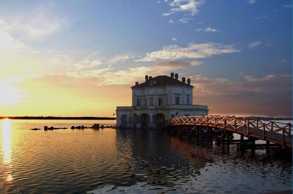 Una villa blanca junto a un lago está conectada por un puente de madera al atardecer.