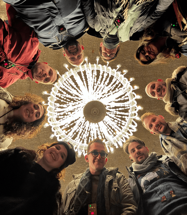 Un gruppo WeRoad con persone sorridenti in cerchio, visto dal basso, con un grande lampadario illuminato al centro.