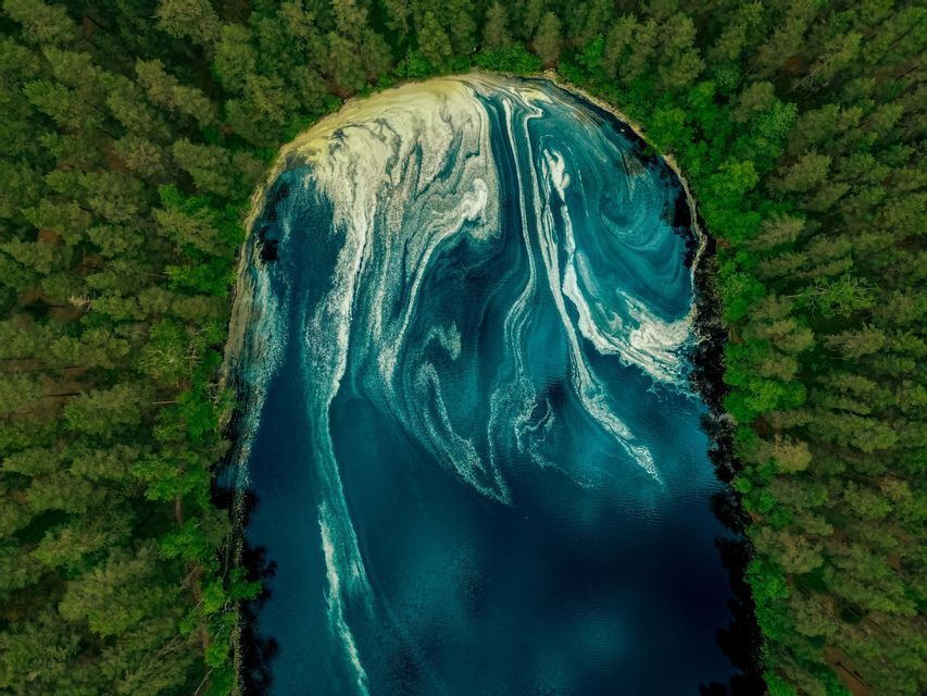 Vista aerea di un lago blu scuro, con motivi bianchi e gialli vorticosi sulla sua superficie, circondato da una fitta foresta verde.