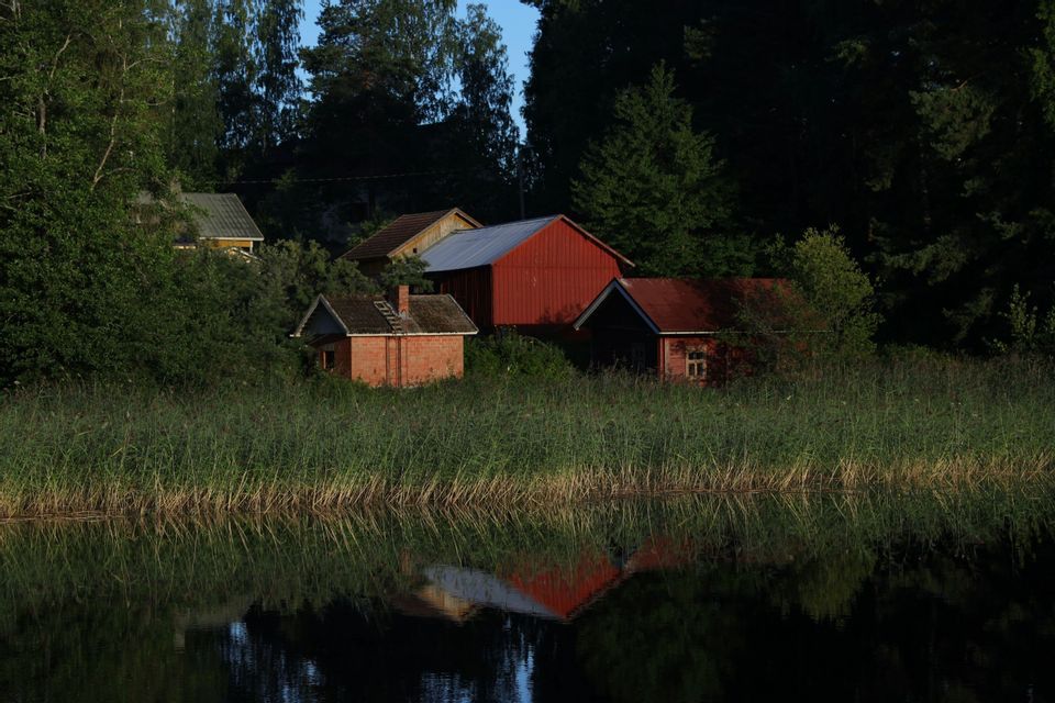 Diversi edifici rustici, tra cui un fienile rosso, sono annidati tra gli alberi sulla riva di un lago e si riflettono nelle calme acque sottostanti.