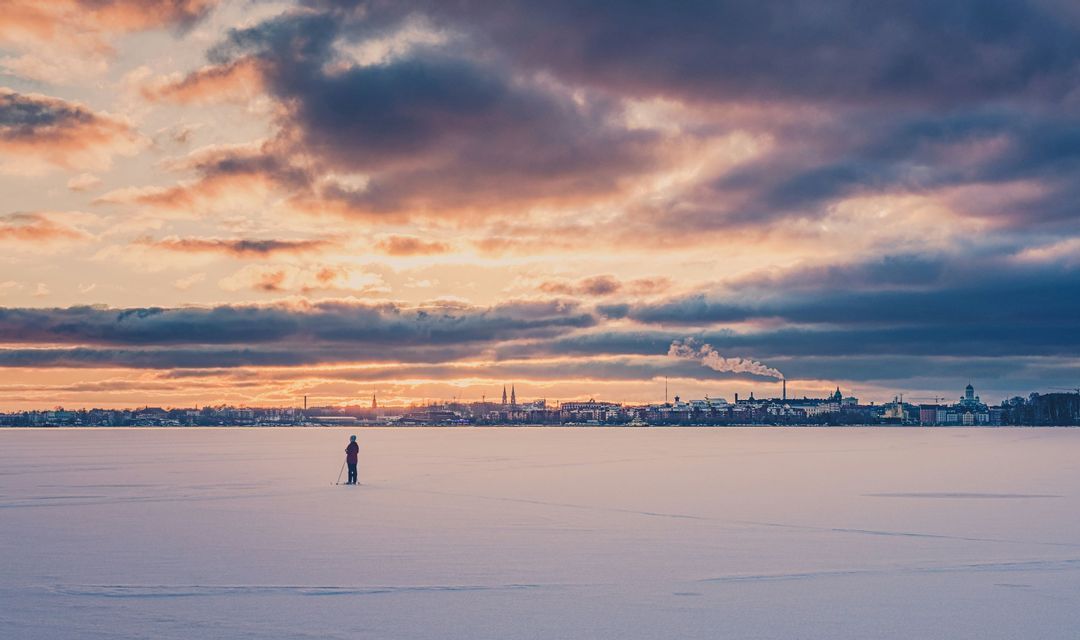 Una persona solitaria si trova su una vasta pianura innevata, osservando lo skyline di una città sotto un drammatico cielo al tramonto.
