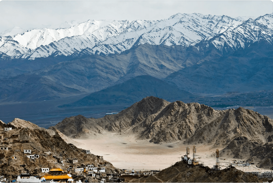 A hillside village overlooks a barren valley with a vast, snow-capped mountain range in the background.