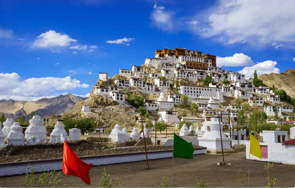 A white monastery complex built into a rocky hillside under a blue sky, with white stupas and colorful flags in the foreground.