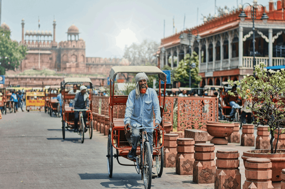 An elderly man with a turban rides a red cycle rickshaw on a busy street, with a historic fort visible in the background.