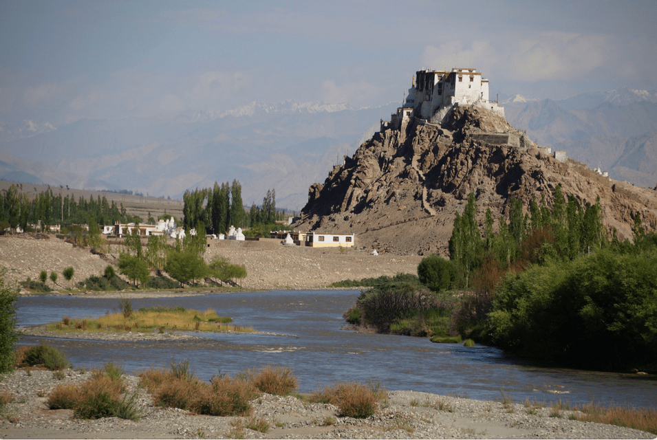 A white monastery sits on a rocky hill overlooking a river valley, with distant hazy mountains in the background.