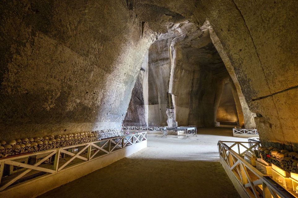Filas de cráneos y huesos humanos se exhiben detrás de barandales blancos dentro de una vasta y luminosa catacumba de piedra con altos techos abovedados.