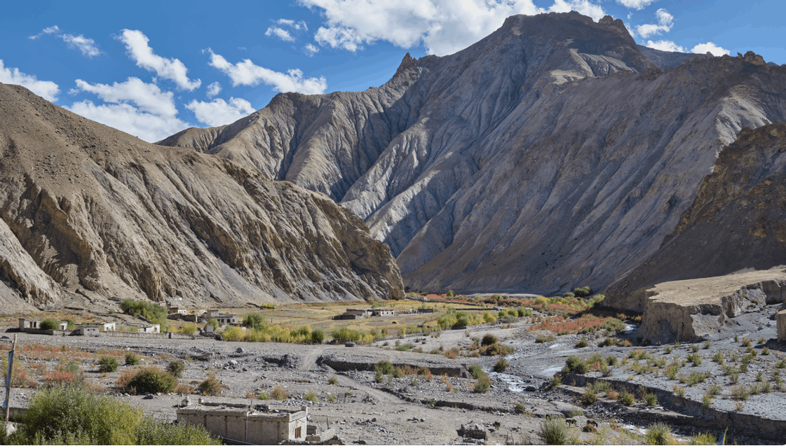 A small village sits in a wide, arid valley at the base of large, rocky mountains under a partly cloudy blue sky.