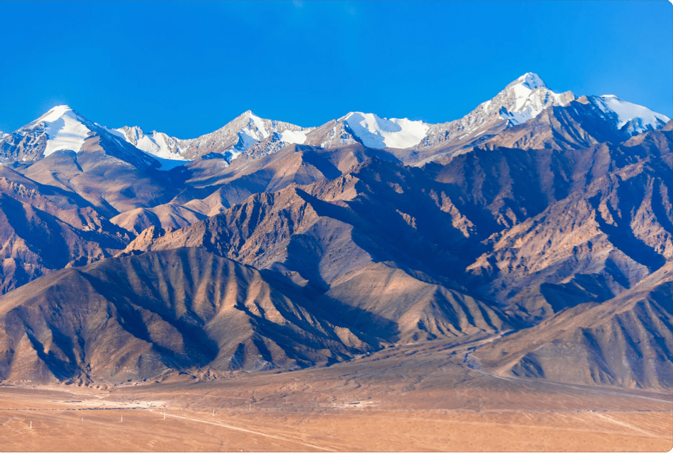 A rugged mountain range with snow-capped peaks stands against a clear blue sky, with a vast desert landscape in the foreground.