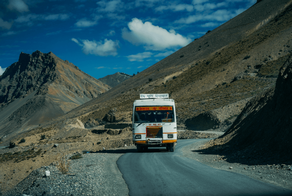 A white bus drives on a winding asphalt road through a rocky, mountainous landscape under a bright blue sky.