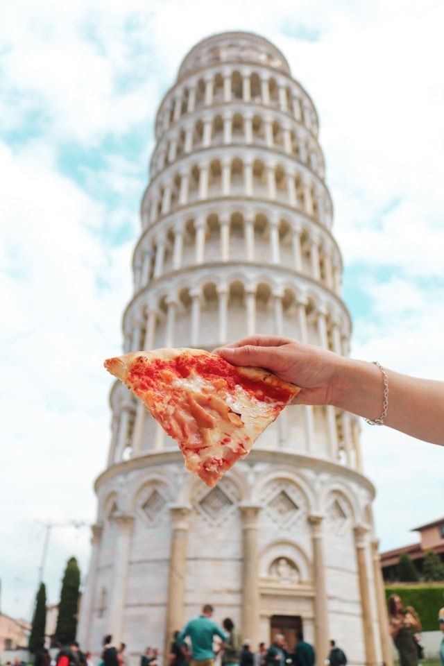 Eine Hand hält ein Stück Käsepizza im Vordergrund, mit dem unscharfen Schiefen Turm von Pisa im Hintergrund.