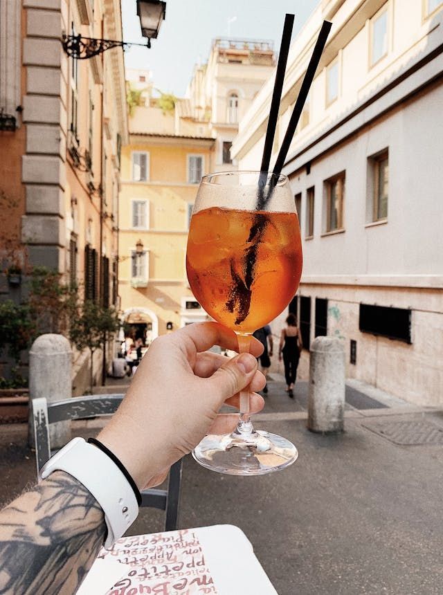 Eine Hand hält ein Glas eines orangefarbenen Cocktails mit zwei Strohhalmen auf einer engen europäischen Stadtstraße.