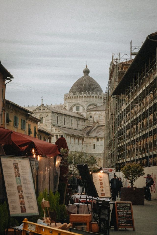 Blick auf eine enge Stadtstraße mit einer großen Kuppelkathedrale, Außengastronomie und einem Gebäude unter Gerüsten im Vordergrund.