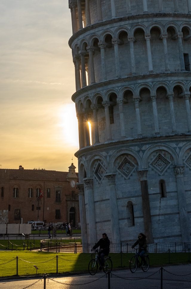 Zwei Personen radeln bei Sonnenuntergang an einem schiefen Steinturm vorbei, wobei die Sonne durch dessen Bögen scheint.