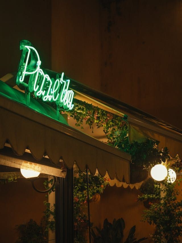 Ein hellgrünes Neonschild mit der Aufschrift 'Pizzeria' leuchtet nachts über einer geschwungenen Markise und Hängepflanzen eines Restaurants.