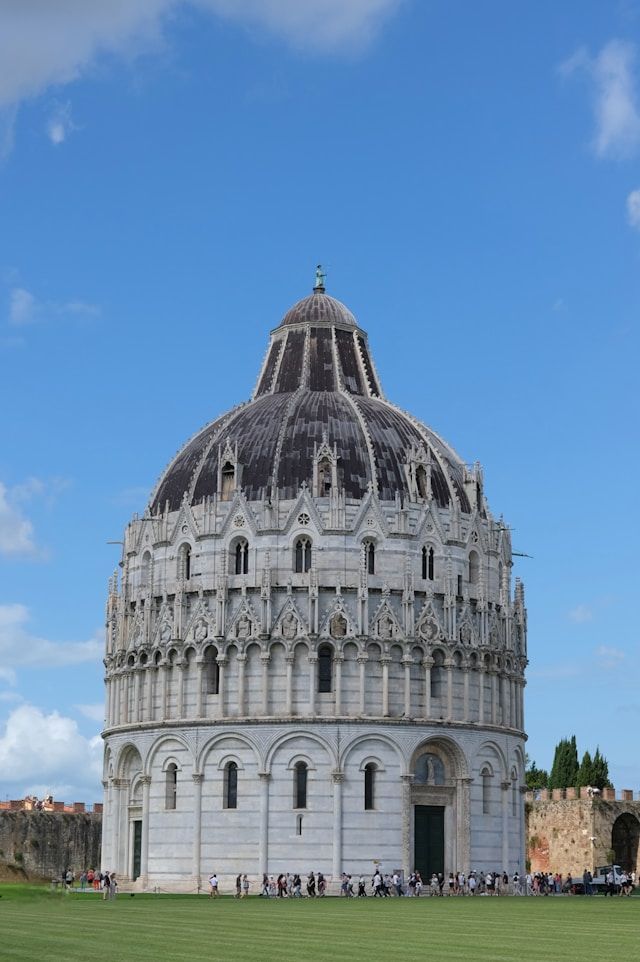 Ein prächtiges Baptisterium aus weißem Marmor mit großer Kuppel steht auf einem grünen Rasen, Menschen versammeln sich an seinem Fuß unter blauem Himmel.