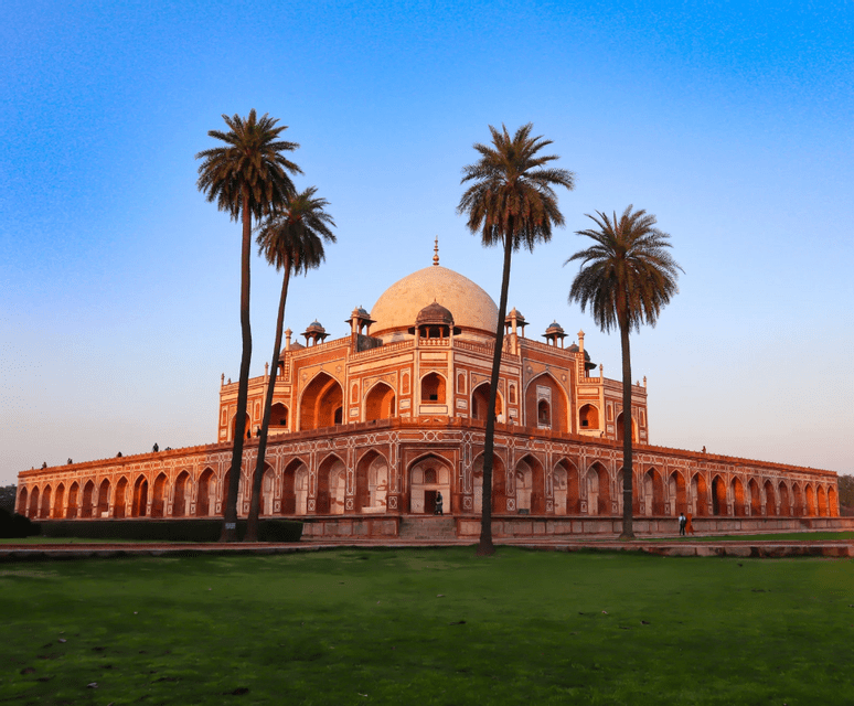 A red and white stone mausoleum with a central dome stands behind a green lawn, with four tall palm trees in front under a clear sky.