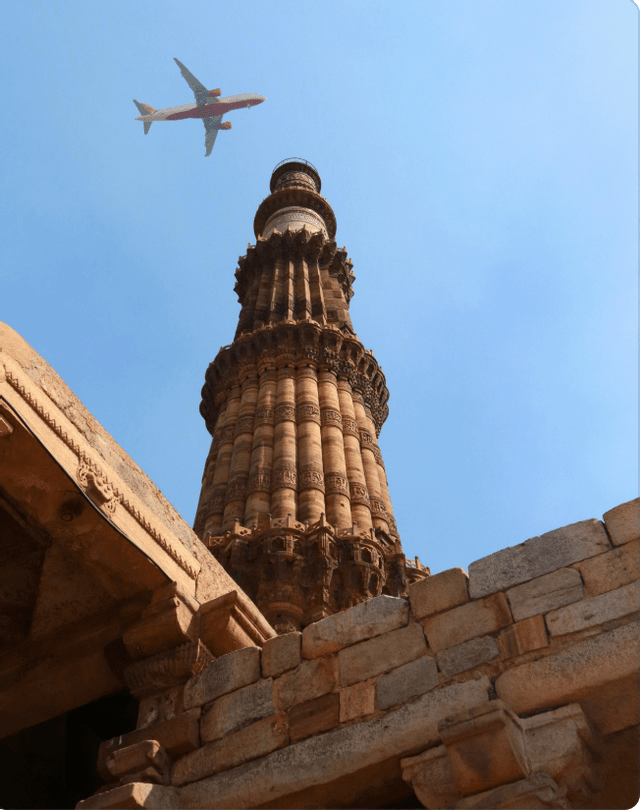 A low-angle view of an airplane flying over a tall, ornate sandstone tower against a clear blue sky, with ancient stone walls in the foreground.