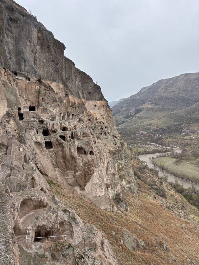 Une cité rupestre taillée dans une falaise, dominant une rivière sinueuse et une vallée sous un ciel couvert.